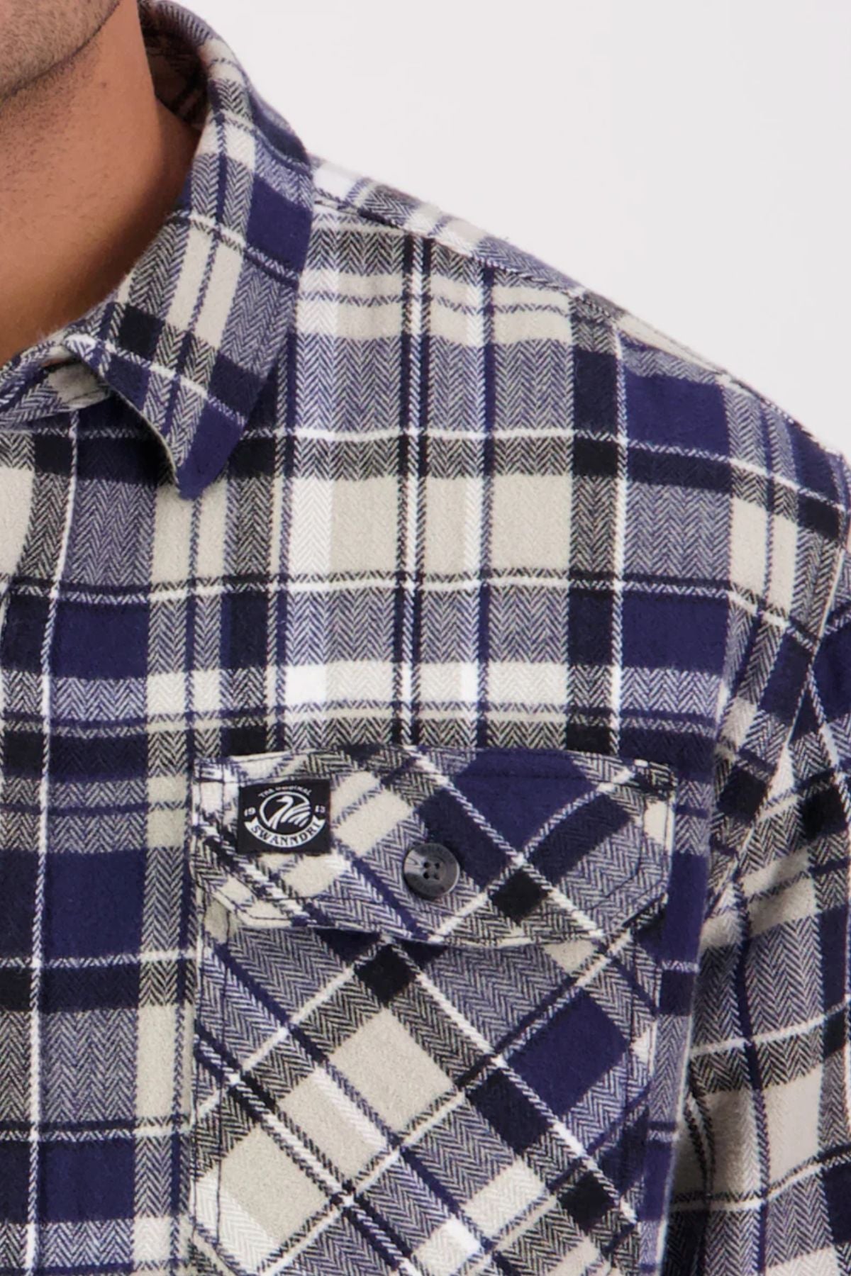 Close-up of a person wearing a Swanndri Morven Sherpa Long Sleeve Shirt in navy, white, and black plaid. Made from 100% cotton flannel with a chest pocket featuring a black button and tag; only neck and shoulder are shown.