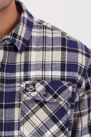 Close-up of a person wearing a Swanndri Morven Sherpa Long Sleeve Shirt in navy, white, and black plaid. Made from 100% cotton flannel with a chest pocket featuring a black button and tag; only neck and shoulder are shown.