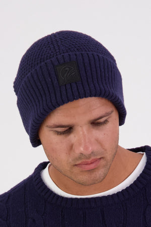 A man wearing the Swanndri Newton Beanie in dark blue and a matching sweater over a white shirt looks downward against a plain light background.