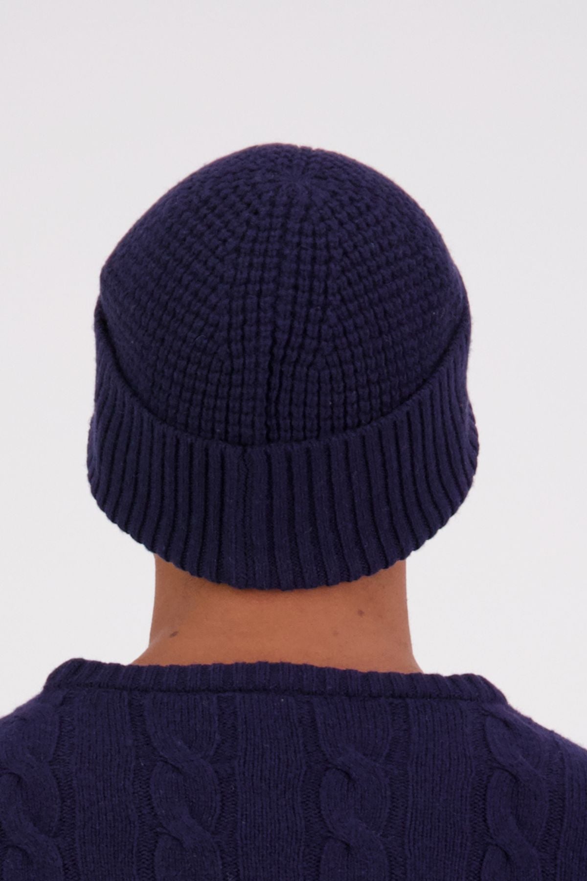 A person is shown from behind against a plain light background, wearing a dark blue Swanndri Newton Beanie and a matching cable-knit sweater.