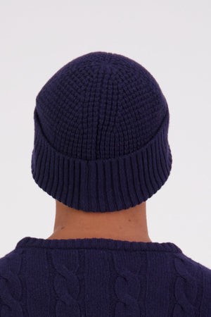 A person is shown from behind against a plain light background, wearing a dark blue Swanndri Newton Beanie and a matching cable-knit sweater.