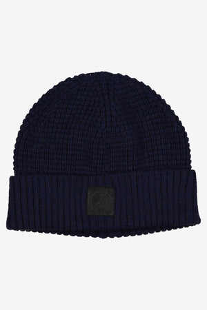 The Swanndri Newton Beanie is a dark blue waffle knit hat with a folded brim and a small black rectangular patch on the front, shown laid flat on a plain white background.