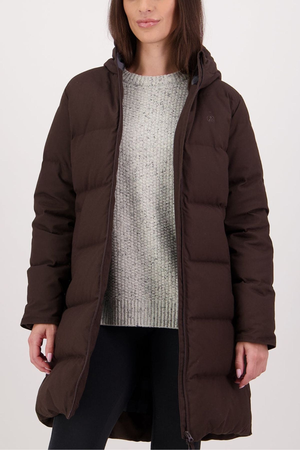 A woman wears the Swanndri Nimbus Longline Down Coat, a long dark brown insulated jacket, over a light gray knit sweater and black pants, standing against a plain light background.