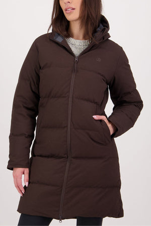 A woman stands against a plain light background, one hand in her pocket, wearing the Swanndri Nimbus Longline Down Coat—long, dark brown, featuring a hood and front zipper. Her face is partially visible.