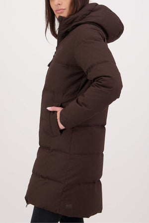 A woman stands in profile, hands in pockets, wearing the Swanndri Nimbus Longline Down Coat—an insulated long, dark brown hooded jacket—against a plain light background.