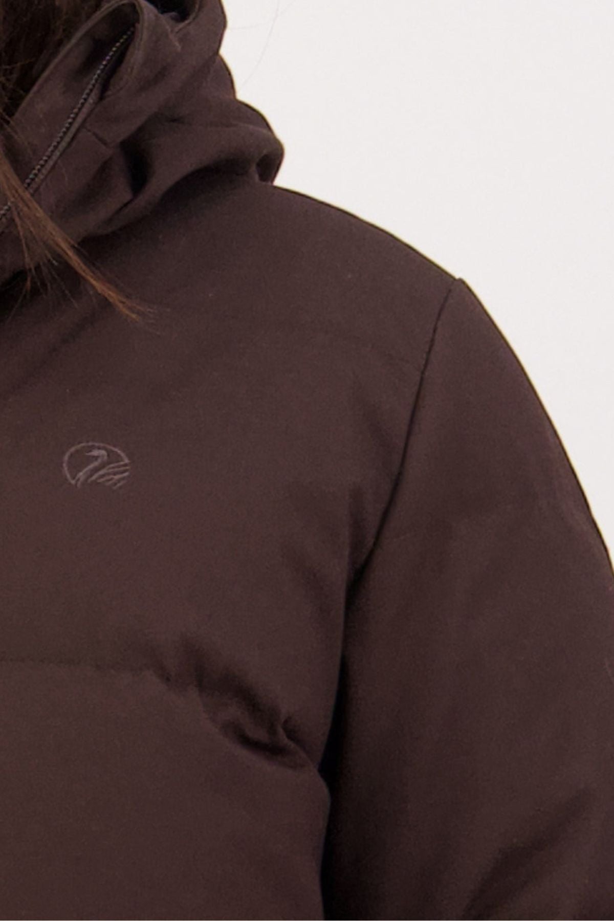 Close-up of a person wearing a dark brown Swanndri Nimbus Longline Down Coat, highlighting the shoulder, chest, and part of the hood with a small light-colored embroidered logo on the chest.