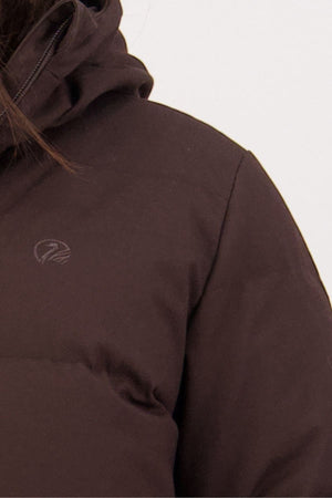 Close-up of a person wearing a dark brown Swanndri Nimbus Longline Down Coat, highlighting the shoulder, chest, and part of the hood with a small light-colored embroidered logo on the chest.
