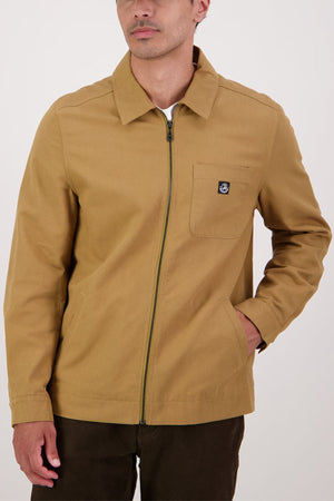 A man wearing the Swanndri Northgate Canvas Jacket in tan, featuring a small black logo patch on the left chest pocket, is paired with dark brown pants and stands with one hand in his pocket against a plain background.