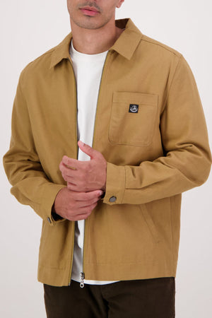A person wears the Swanndri Northgate Canvas Jacket in tan over a white t-shirt, touching their chest. The jacket features a chest pocket with a small black logo patch and buttoned cuffs, set against a plain light background.