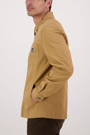 A person wearing the Swanndri Northgate Canvas Jacket in tan, with one hand in the pocket, is photographed from the side against a plain light background.