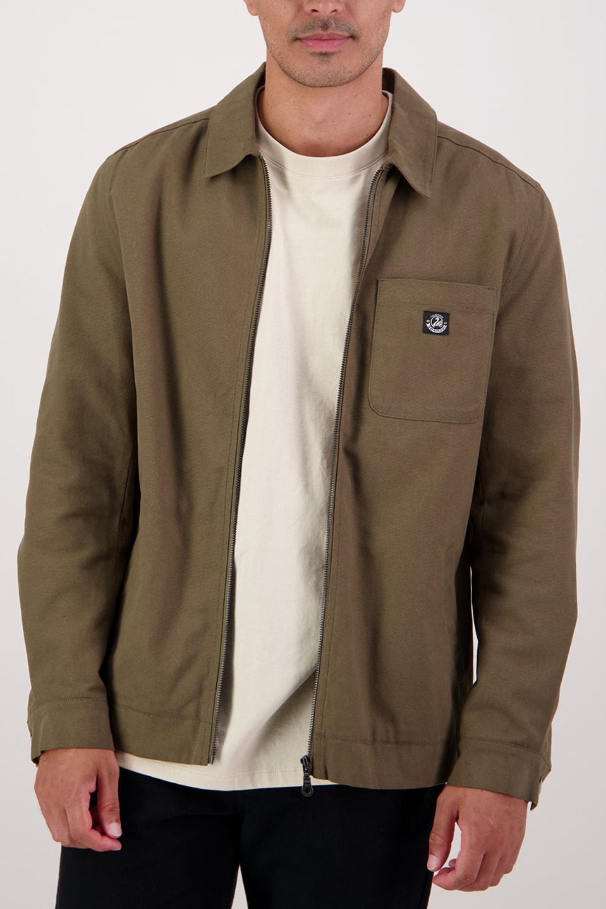 A person wears the Swanndri Northgate Canvas Jacket in olive green with a chest pocket, layered over a cream t-shirt; only their upper body is shown against a plain white background.