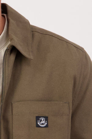 A close-up of a person wearing the Swanndri Northgate Canvas Jacket in olive green with a zip-up front and chest pocket featuring a small rectangular patch with a circular logo, set against a plain light background.