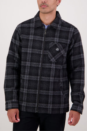 A man stands against a plain light background wearing the Swanndri Northgate V2 Jacket, featuring a black and gray plaid pattern, a zip-up front, chest pocket, and paired with black pants.