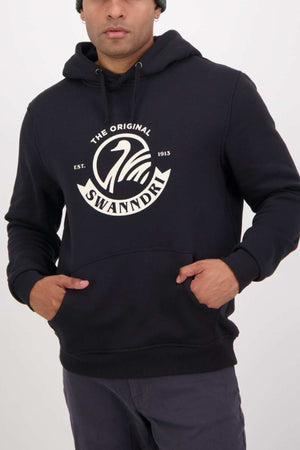 A person sports the iconic Swanndri Original V2 Hoodie in black, decorated with a stylish white logo featuring a swan and "The Original Swanndri Est. 1913" text. They exude timeless style with hands nestled in the kangaroo pocket.