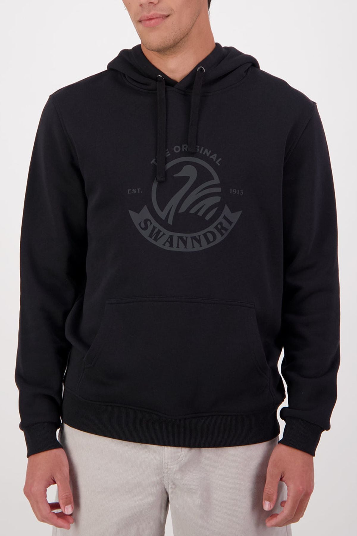 Someone wears the Swanndri Original V2 Hoodie—a black cotton-polyester hoodie featuring a kangaroo pocket and a subtle Swanndri logo with swan graphic on the chest—paired with light-colored pants.