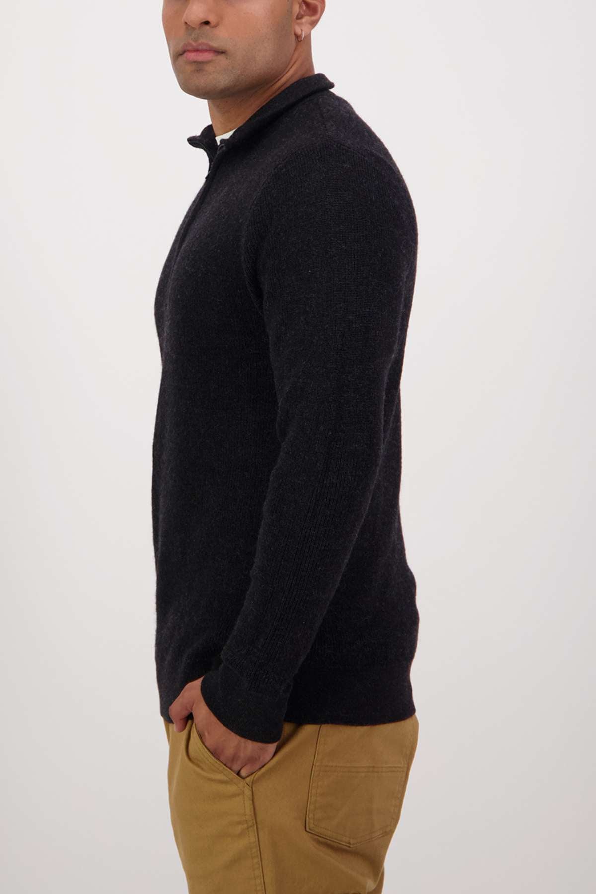 A person wearing a Swanndri Otahu River Quarter Zip Merino Knit in dark color and tan pants stands in profile against a plain background. Their hands are in pockets, and the sweater features a high collar.