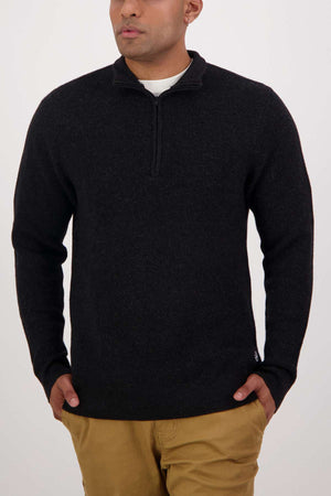 A person wearing the Swanndri Otahu River Quarter Zip Merino Knit in black and tan pants stands against a plain white backdrop, hands in pockets.