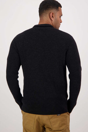 A person with short hair, wearing a Swanndri Otahu River Quarter Zip Merino Knit sweater and tan pants, faces away. The plain, neutral background highlights their clothing.