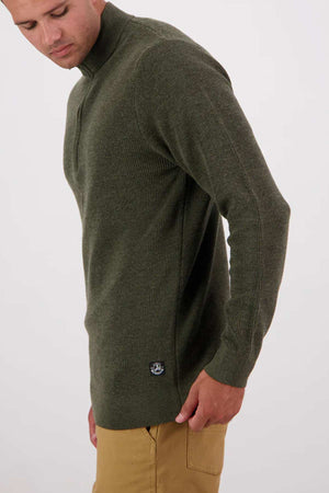 A person wearing an olive green Swanndri Otahu River Quarter Zip Merino Knit with tan pants, standing against a plain white background.