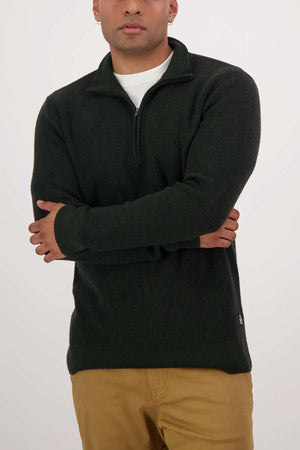 A person in a dark green Swanndri Otahu River Quarter Zip Merino Knit over a white shirt and mustard-colored pants stands with arms crossed against a plain light background.