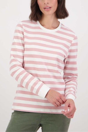 A woman models the Swanndri Papamoa Stripe Long Sleeve Tee in pink and white with white cuffs and a relaxed fit, paired with olive green pants. She stands against a plain background, her brown hair framing her face—a versatile look for everyday wear.