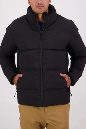 A man stands against a plain light background, hands in his pockets, wearing tan pants and the Swanndri Plume Mens Down Jacket with duck down fill, high collar, and zipper pockets—ideal for outdoor adventures.