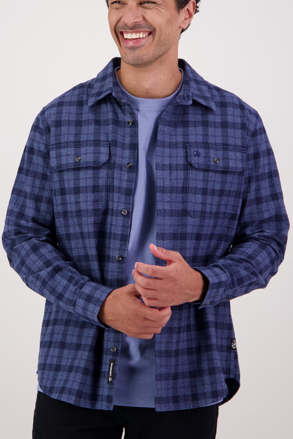 A smiling man stands with his hands clasped in front of him against a plain light background, wearing the Swanndri Port Road Shirt in blue and black heavyweight twill over a light blue t-shirt.