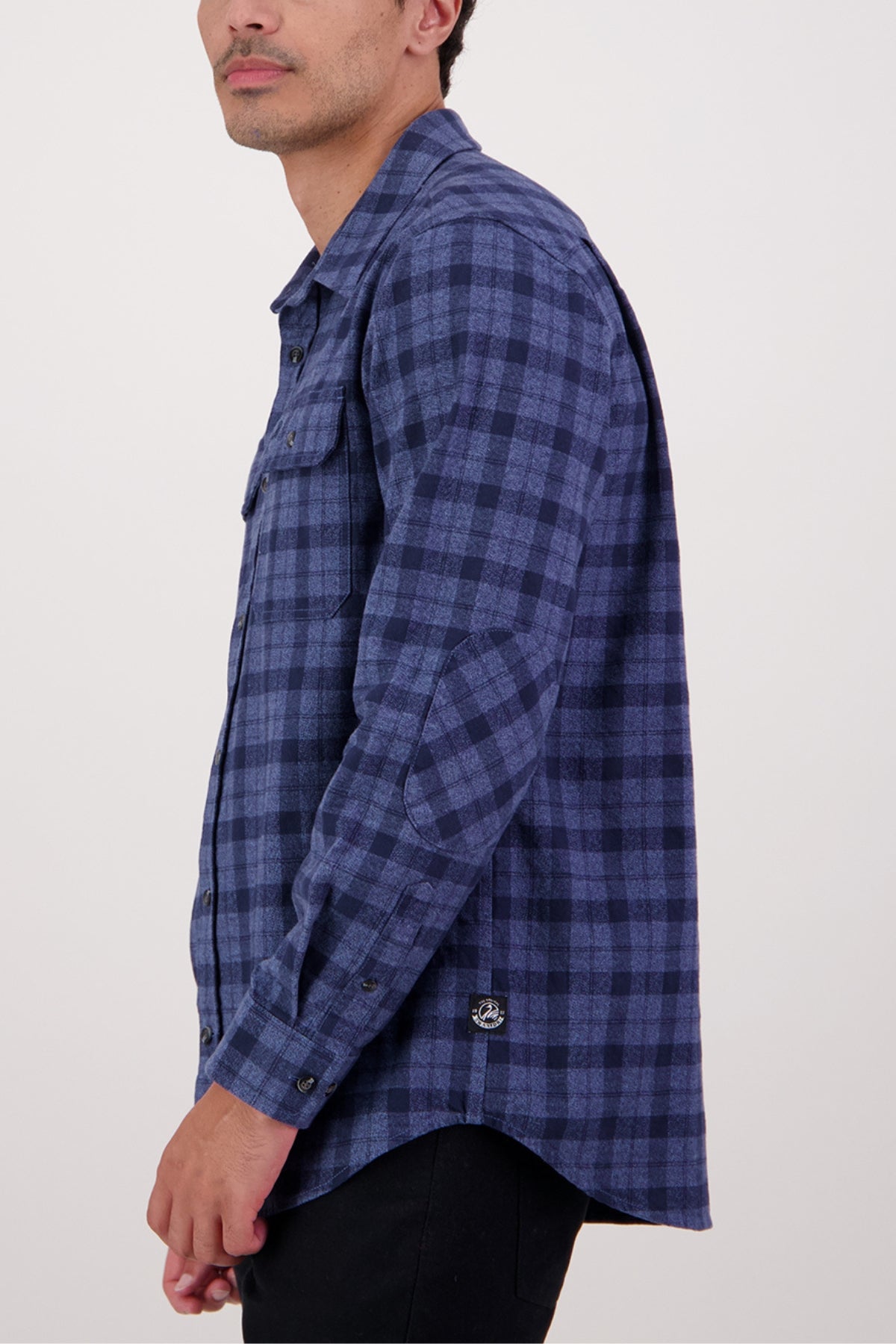 A person wearing a Swanndri Port Road Shirt in blue and black plaid heavy weight twill, with long sleeves and chest pockets, stands sideways facing left. The 100% cotton shirt is paired with black pants.