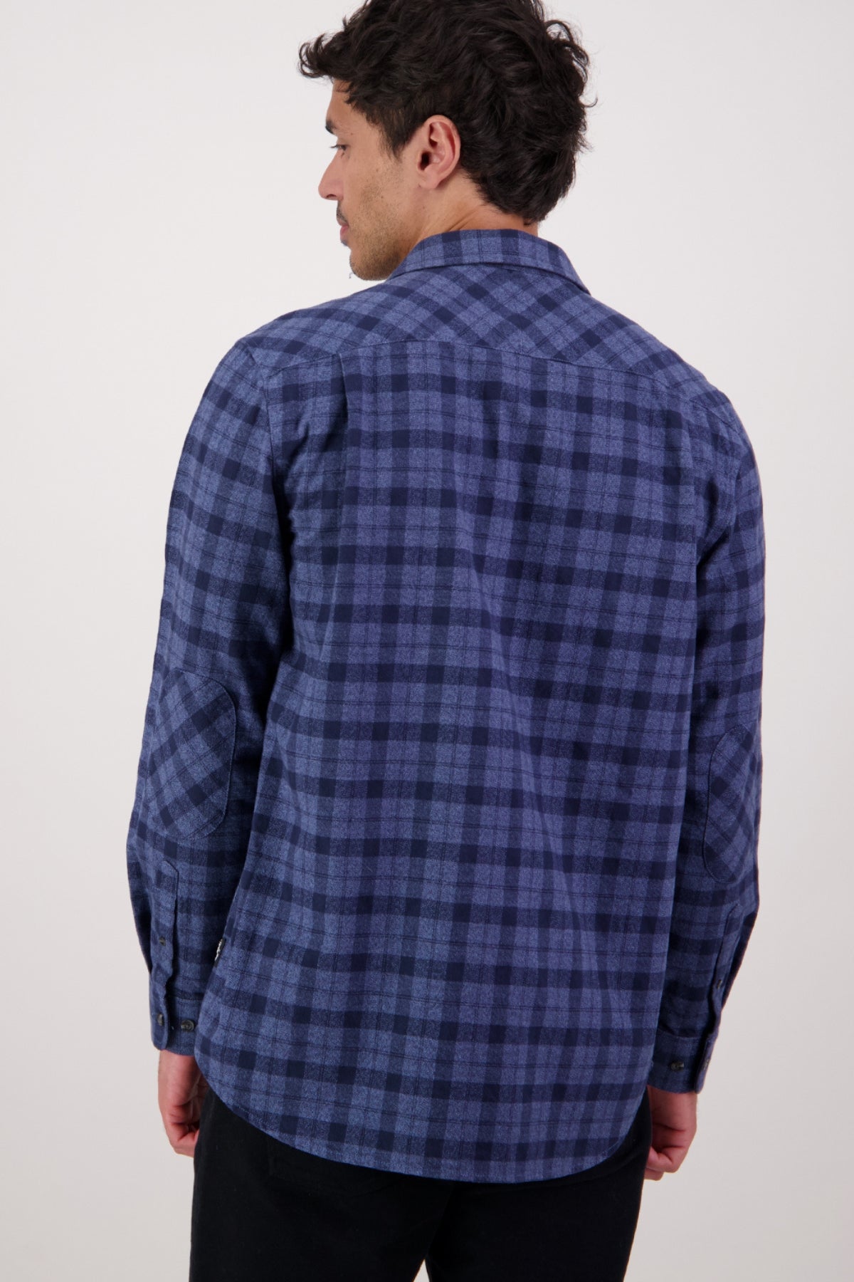 A man with short dark hair is seen from behind, wearing a Swanndri Port Road Shirt in blue and navy checks, made from 100% cotton, paired with black pants against a plain light-colored background.