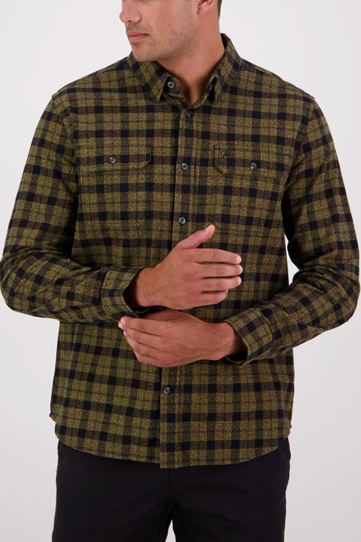 A man stands against a plain background, looking to the side and touching his chest, wearing the Swanndri Port Road Shirt in green and black plaid, made from heavyweight cotton twill with two chest pockets.