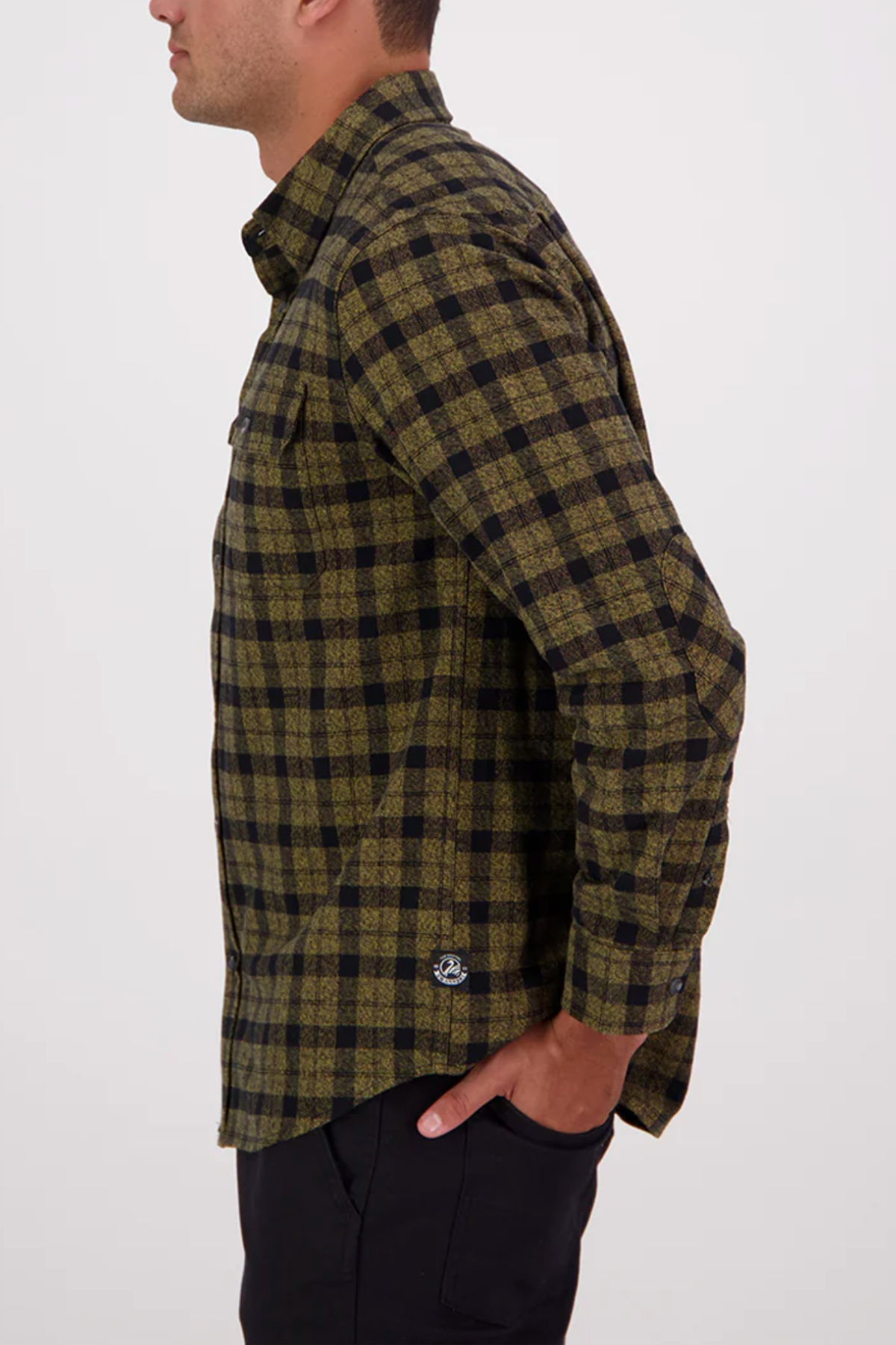A person stands in profile against a plain background, wearing the Swanndri Port Road Shirt with a green and black check pattern and black pants, one hand in their pocket. The shirt is made from heavyweight cotton twill.