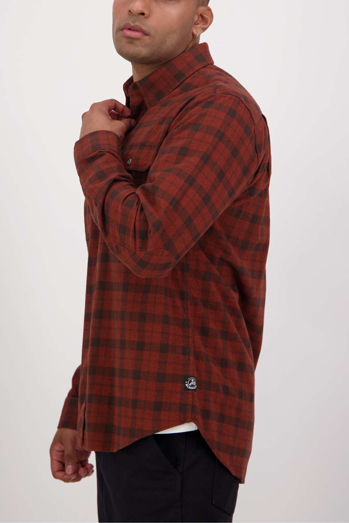 A person wearing a Swanndri Port Road Shirt, featuring a red and black checkered cotton design with buttoned chest pockets, stands sideways adjusting the collar. The shirt includes a classic button-up style and a small logo at the hem against a plain white background.