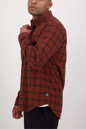 A person wearing a Swanndri Port Road Shirt, featuring a red and black checkered cotton design with buttoned chest pockets, stands sideways adjusting the collar. The shirt includes a classic button-up style and a small logo at the hem against a plain white background.