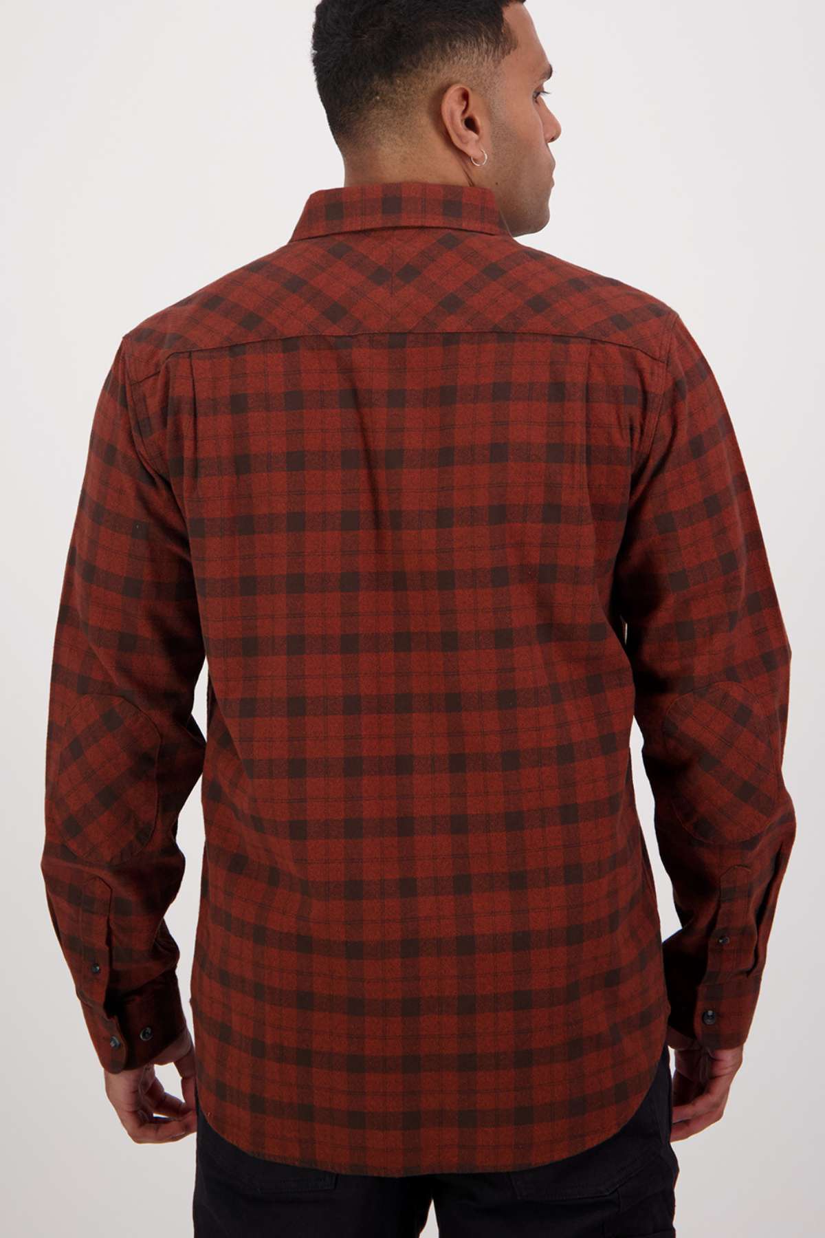 A person faces away from the camera wearing a Swanndri Port Road Shirt, a long-sleeved red and black checkered cotton shirt with buttoned chest pockets, a collar, and buttoned cuffs. It is perfectly paired with sleek black pants.