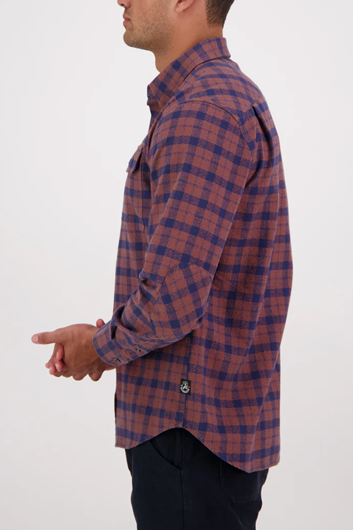 A man with dark hair clasps his hands together while wearing the Swanndri Port Road Shirt, shown from the side against a plain light background. The shirt features a brown and navy check pattern in cotton twill.