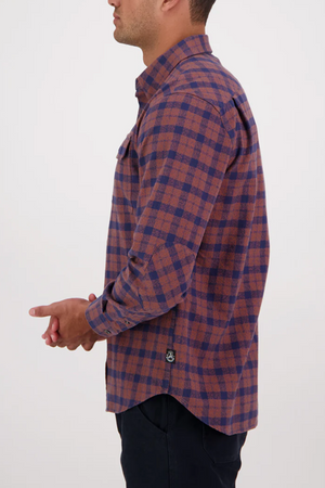 A man with dark hair clasps his hands together while wearing the Swanndri Port Road Shirt, shown from the side against a plain light background. The shirt features a brown and navy check pattern in cotton twill.