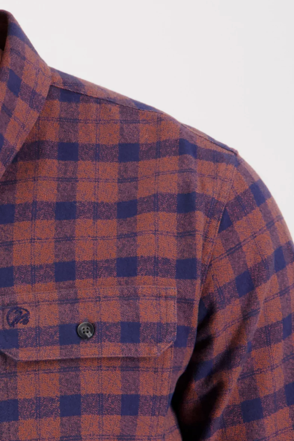 A close-up of the Swanndri Port Road Shirt shows its yarn-dyed check cotton twill in dark blue and rust-orange, a buttoned chest pocket, and part of the collar against a light background.