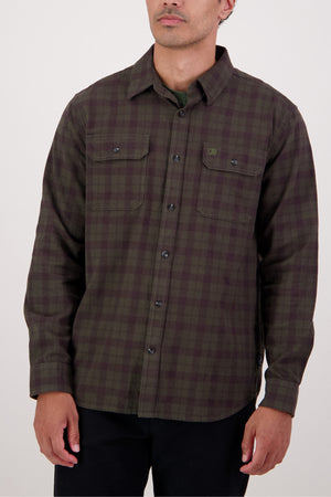 A person wearing the Swanndri Port Road Shirt—a heavyweight twill shirt in dark green and brown plaid with two chest pockets—stands against a plain light background.