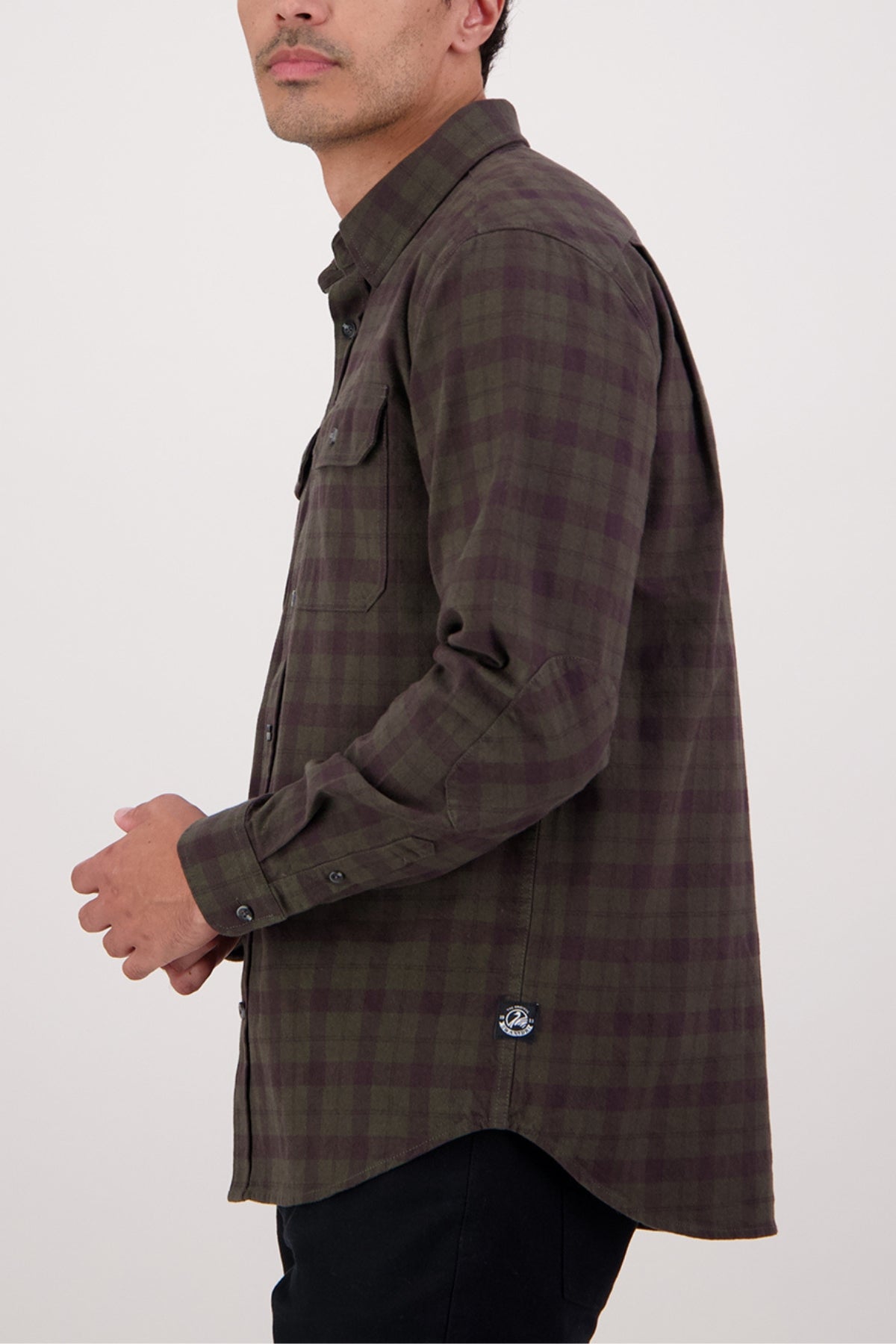 A man stands sideways against a plain light background, wearing the Swanndri Port Road Shirt—a heavyweight twill with a brown and green plaid pattern, chest pockets, and a small logo tag near the hem.