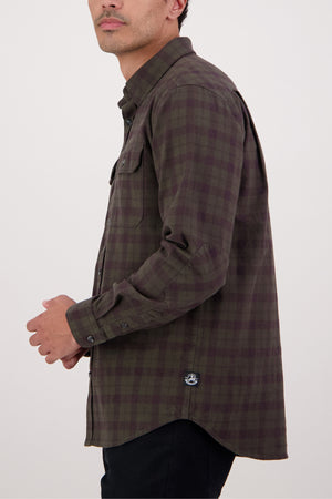 A man stands sideways against a plain light background, wearing the Swanndri Port Road Shirt—a heavyweight twill with a brown and green plaid pattern, chest pockets, and a small logo tag near the hem.