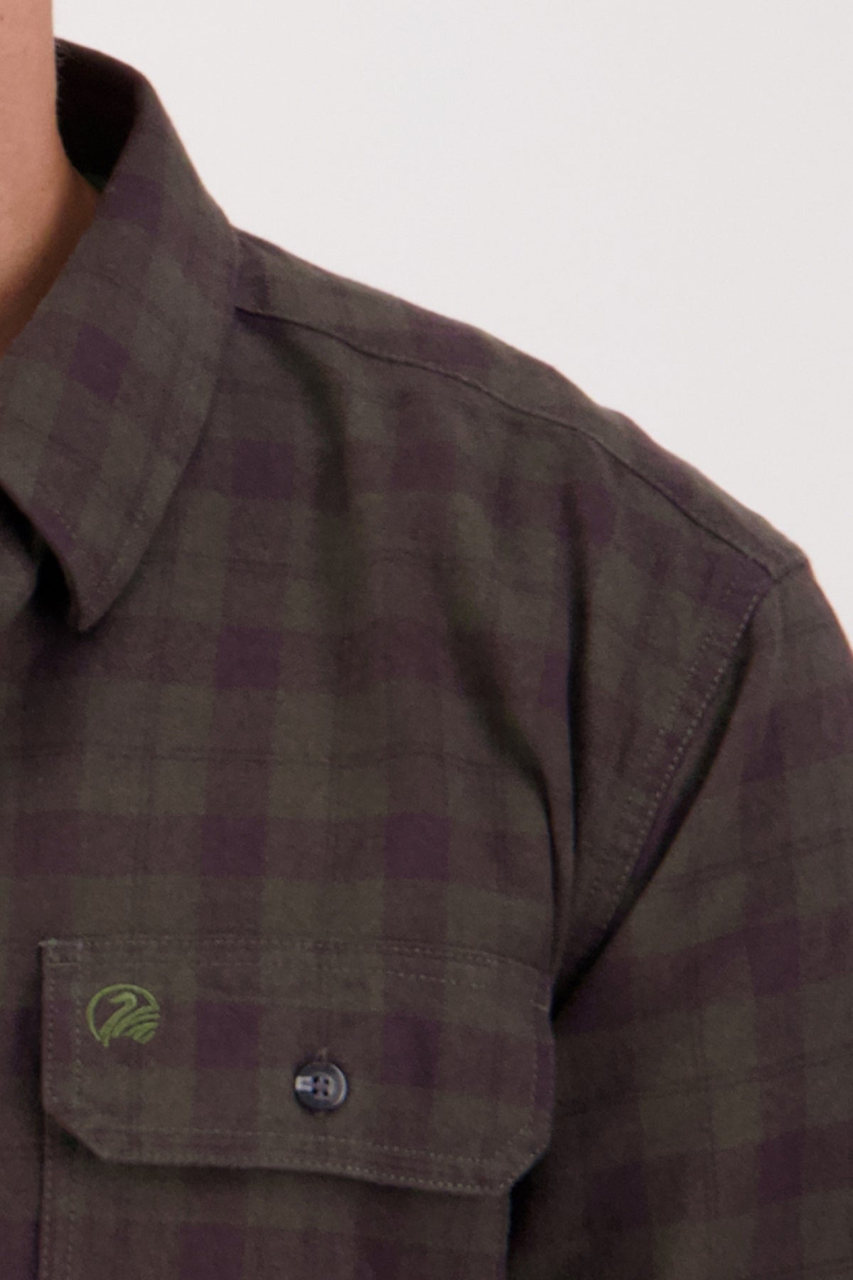 A close-up of a person wearing the Swanndri Port Road Shirt in green and purple plaid, highlighting the collar, shoulder, and chest pocket against a plain light background.