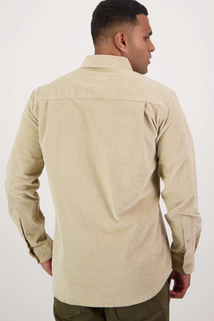 A person with short dark hair is standing with their back to the camera, wearing a Swanndri Ranfurly Corduroy Shirt in light beige. The heavy-weight cotton shirt has a long-sleeve, button-up design, and features a collar and cuffs with a tailored fit. The individual is also wearing green pants against a plain white background.