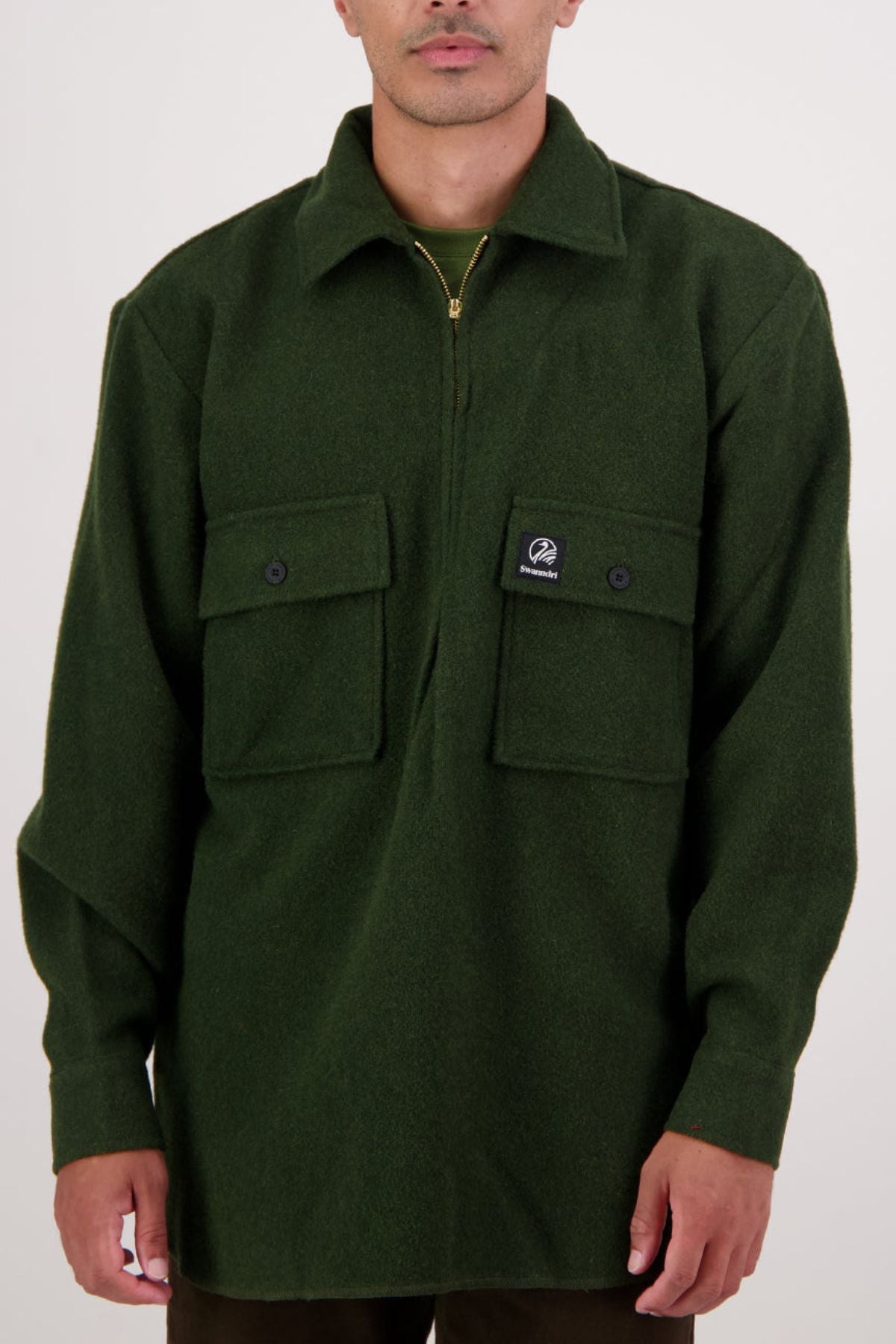 A person wearing a forest green Swanndri Ranger Bush Shirt with two buttoned chest pockets and a small black logo patch on the left pocket stands against a plain light background.