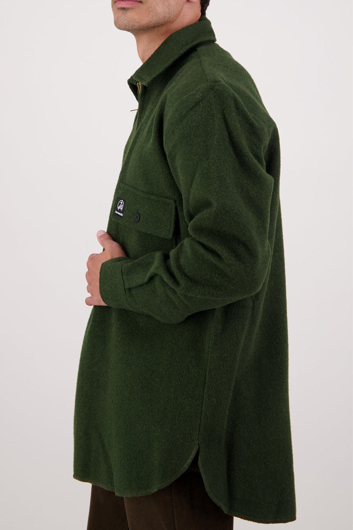 A person wearing an oversized Swanndri Ranger Bush Shirt with a front pocket stands against a plain light background, shown from shoulders to mid-thigh, facing sideways with their hand near the pocket.