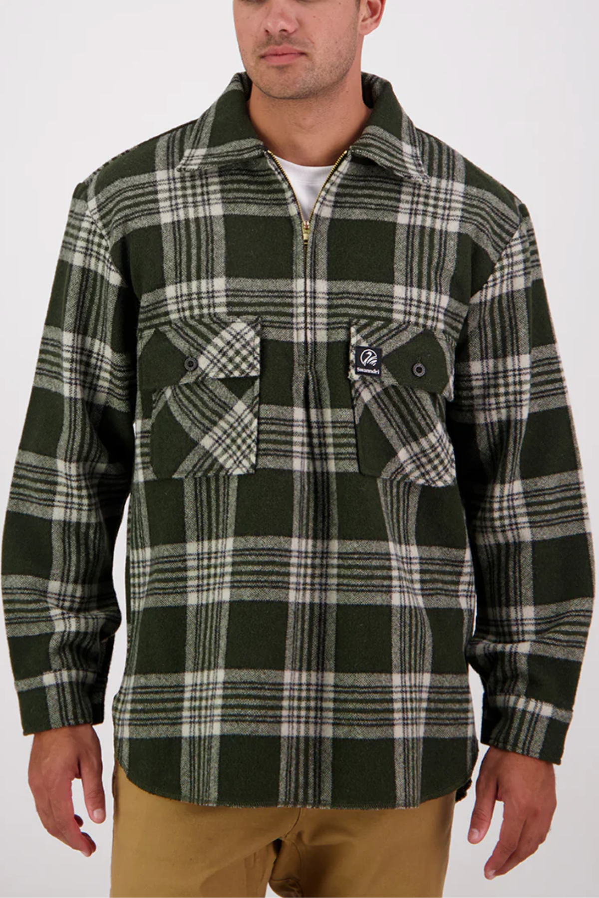 A man stands against a plain white background wearing an oversized green and white plaid zip-up shirt jacket inspired by the Swanndri Ranger Bush Shirt, featuring two chest pockets, paired with tan pants.