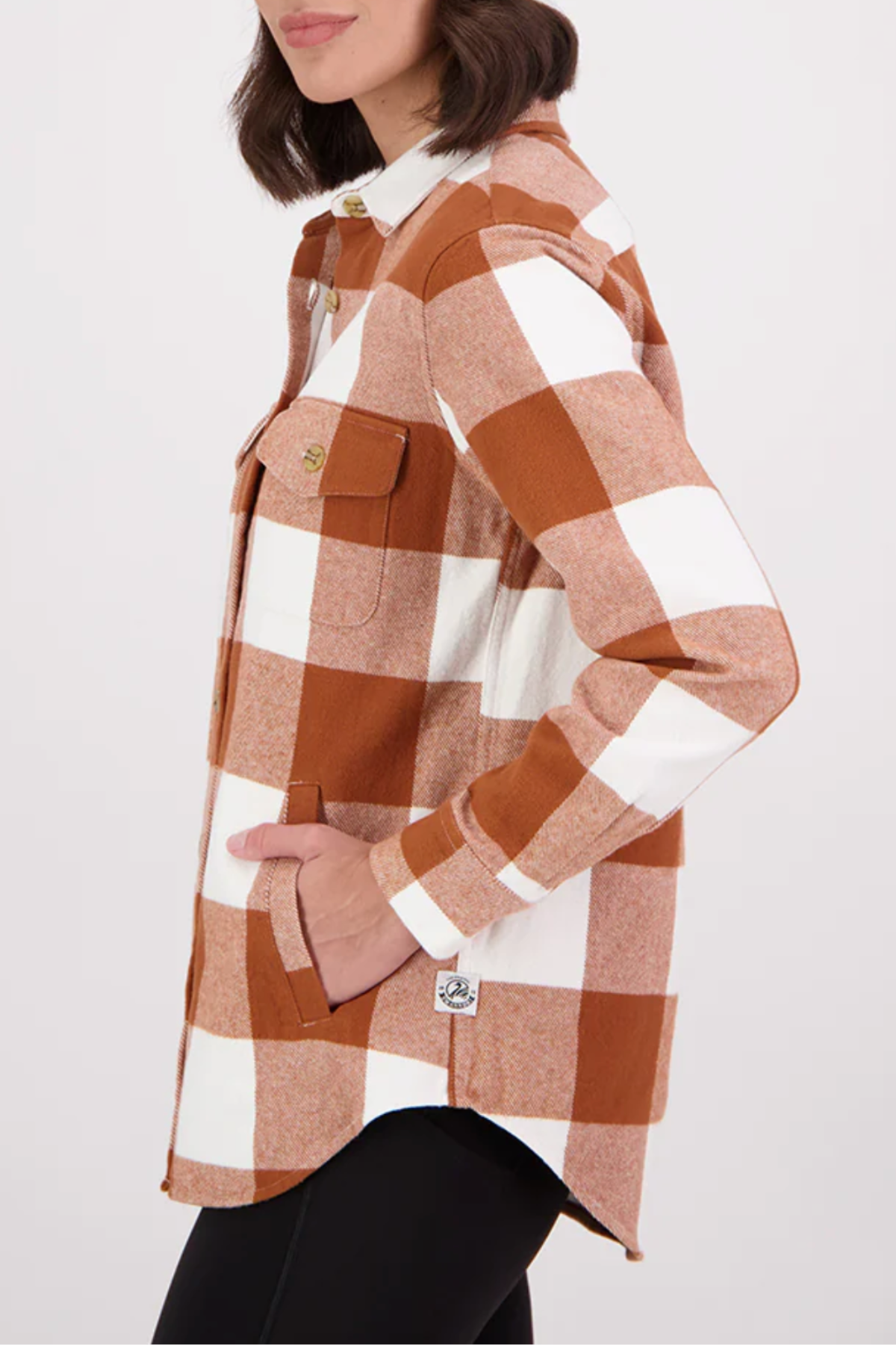 A woman stands in side profile against a plain background, wearing the Swanndri Rapaki Long Sleeve Shacket in brown and white large check, made from 100% brushed cotton. Styled with black pants, it's the perfect winter layer.