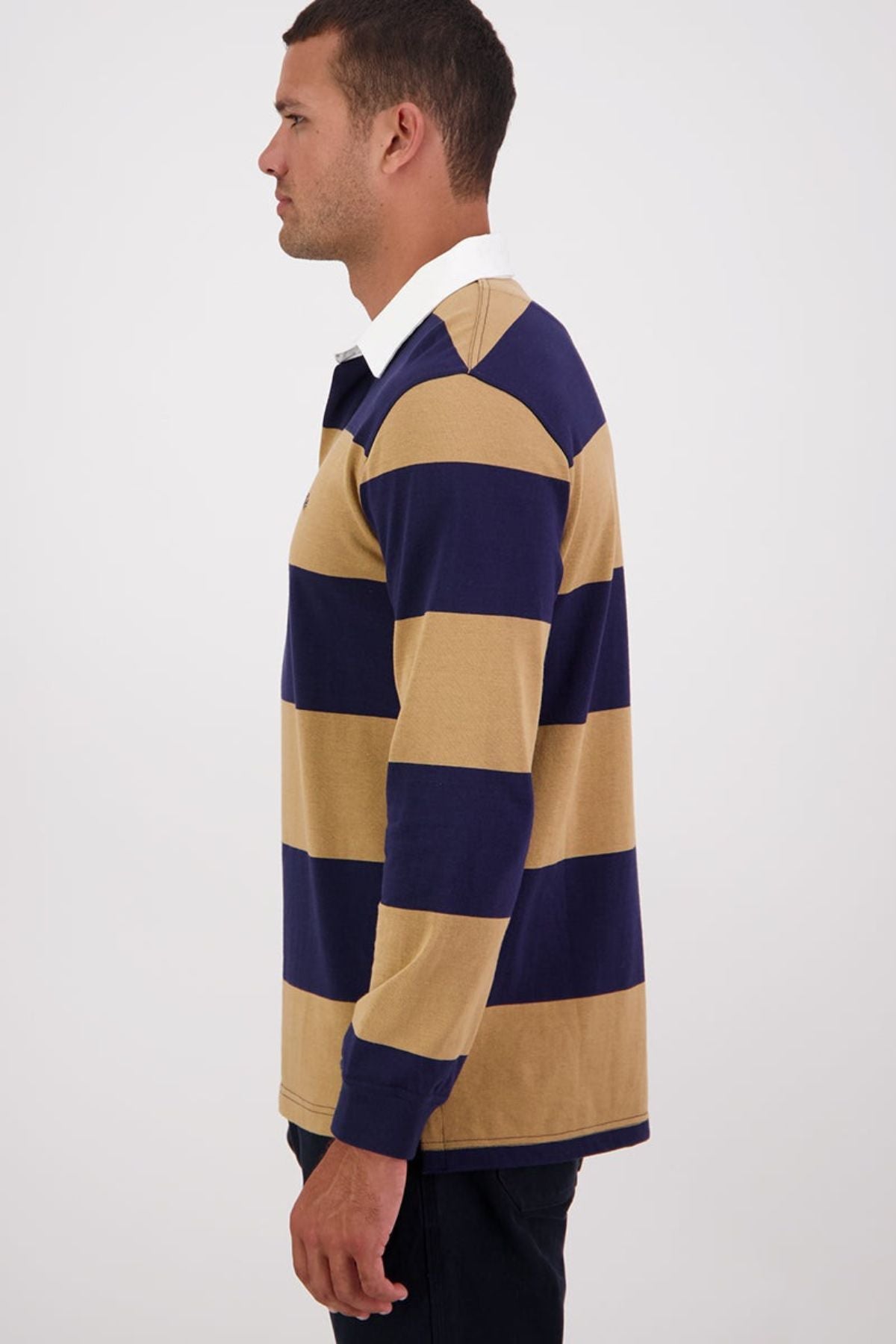 A man in profile wears the Swanndri Ridgeway Rugby, a cotton top with wide navy blue and tan stripes, rubber buttons, and a white collar, standing against a plain light background.