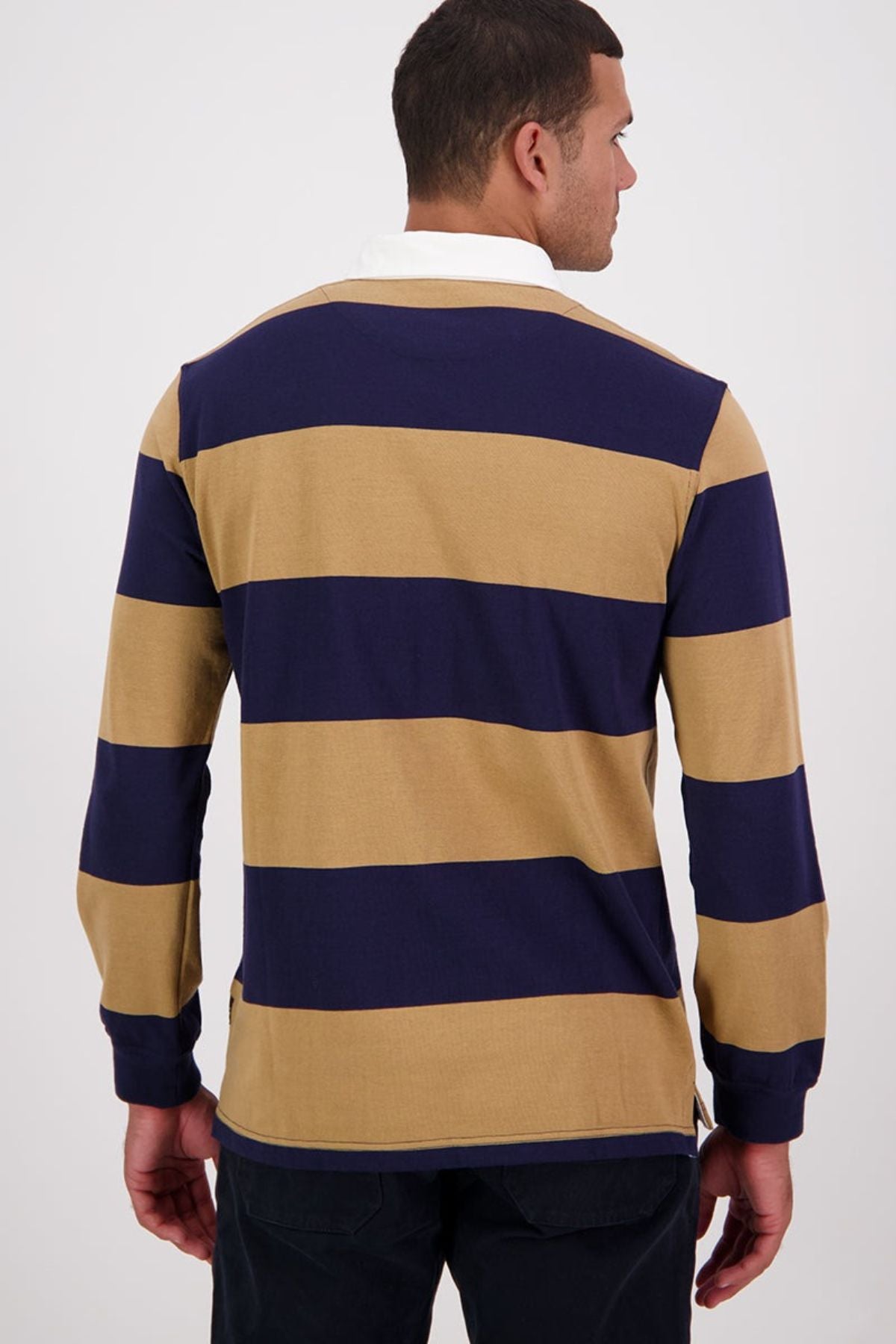 A man is seen from the back wearing the Swanndri Ridgeway Rugby shirt, featuring wide navy blue and tan stripes, a white collar, and Swanndri embroidery, set against a plain light background.