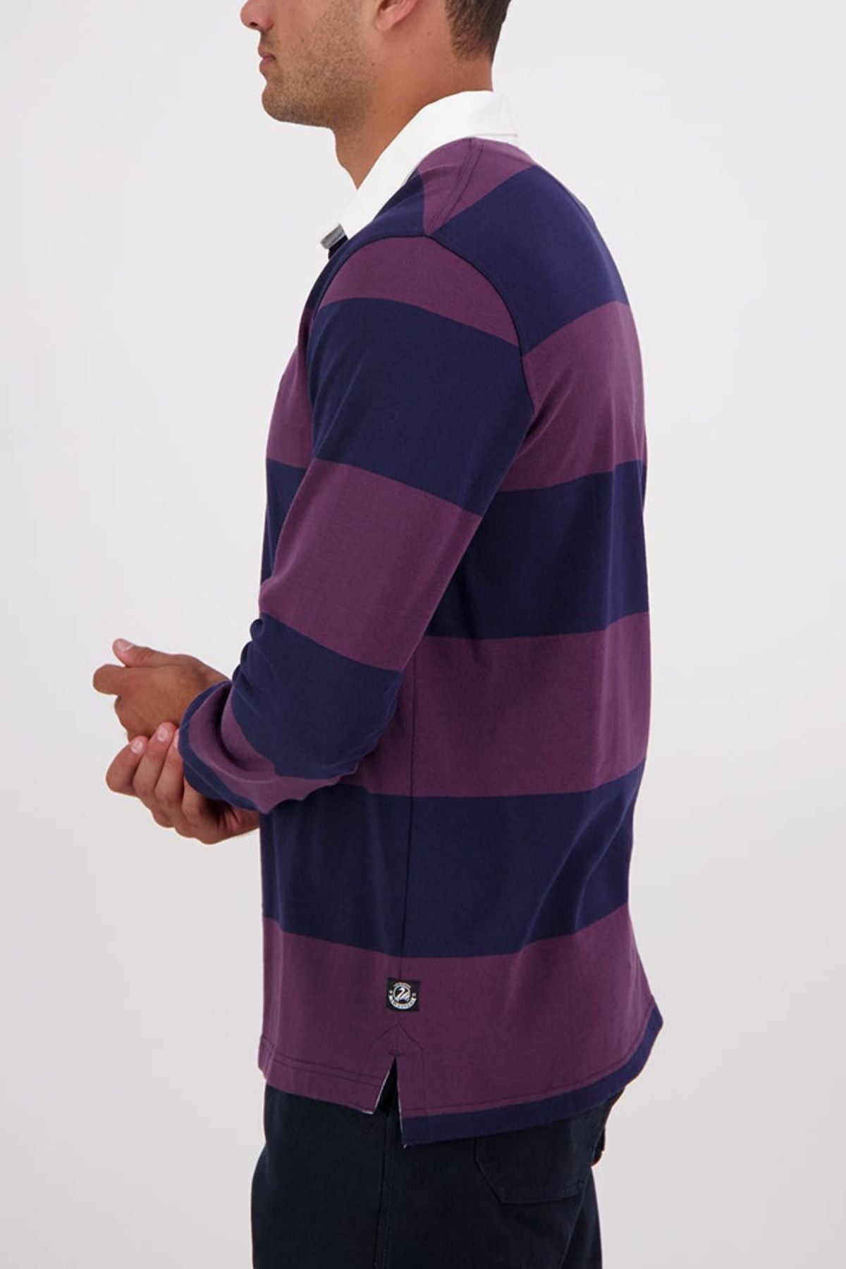 A man stands sideways against a plain white background, hands loosely clasped, wearing the Swanndri Ridgeway Rugby—a long-sleeve, collared shirt with wide purple and navy stripes made from heavy drill cotton.