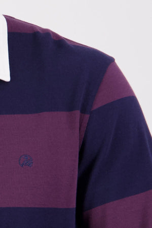 A close-up shows someone wearing the Swanndri Ridgeway Rugby—a purple and dark blue striped shirt with a white collar, made from heavy drill cotton. Only the shoulder and chest are visible.